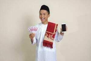 Portrait of attractive Asian muslim man in white shirt with skullcap showing one hundred thousand rupiah while showing blank screen mobile phone. Financial and shopping concept. Isolated image on gray photo