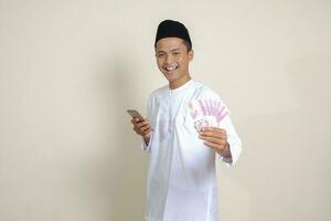 Portrait of attractive Asian muslim man in white shirt showing one hundred thousand rupiah while using mobile phone. Financial and savings concept. Isolated image on gray background photo