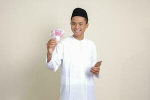 Portrait of attractive Asian muslim man in white shirt showing one hundred thousand rupiah while using mobile phone. Financial and savings concept. Isolated image on gray background photo