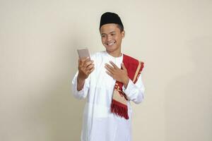 Portrait of attractive Asian muslim man in white shirt taking picture of himself or selfie, saying hi and waving his hand during video call. Isolated image on gray background photo