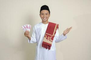 Portrait of attractive Asian muslim man in white shirt showing one hundred thousand rupiah while pointing to the side. Financial and savings concept. Isolated image on gray background photo