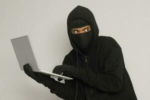 Portrait of mysterious man wearing black hoodie and mask doing hacking activity on laptop, hacker holding a personal computer. Cyber security concept. Isolated image on gray background photo