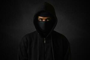 Mysterious man wearing black hoodie and mask standing against dark background, looking at camera. Dramatic low light portrait photo