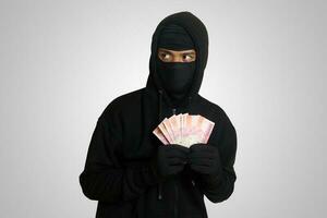 Portrait of mysterious man wearing black hoodie and mask stealing one hundred thousand rupiah successfully from victim. Isolated image on gray background photo