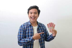Portrait of excited Asian man in blue plaid shirt standing against white background, showing one hundred thousand rupiah while pointing to the side. Financial and savings concept. photo