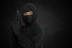 Mysterious man wearing black hoodie and mask holding a pistol, shooting with a gun. Isolated image on dark ambient background photo