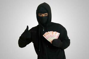 Portrait of mysterious man wearing black hoodie and mask stealing one hundred thousand rupiah successfully from victim. Isolated image on gray background photo