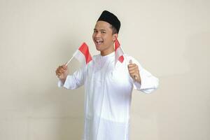 Portrait of attractive Asian muslim man in white shirt with skullcap holding indonesia flag while raising his fist, celebrating success. Isolated image on gray background photo