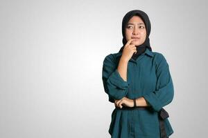 Portrait of confused Asian muslim woman with hijab standing against white background, looking up and thinking about question with hand on chin photo