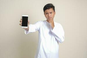 Portrait of attractive Asian muslim man in white shirt showing and presenting blank screen mobile phone. Advertising concept. Isolated image on gray photo