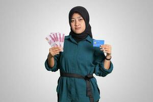 Portrait of cheerful Asian muslim woman with hijab, showing one hundred thousand rupiah while holding a credit card. Financial and savings concept. Isolated image on white background photo