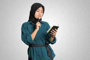 retrato de emocionado asiático musulmán mujer con hijab escritura en Nota libro y pensando con bolígrafo en mentón. publicidad concepto. aislado imagen en blanco antecedentes foto