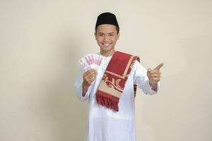 Portrait of attractive Asian muslim man in white shirt showing one hundred thousand rupiah while pointing to the side. Financial and savings concept. Isolated image on gray background photo