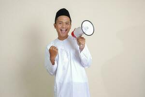 Portrait of attractive Asian muslim man in white shirt with skullcap speaking louder using megaphone, promoting product. Advertising concept. Isolated image on grey background photo