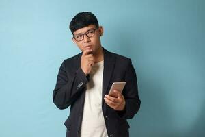 Portrait of young Asian business man in casual suit thingking while holding phone. Isolated image on blue background photo
