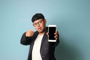 Portrait of young Asian business man in casual suit pointing finger at camera with surprised face while holding phone. Isolated image on blue background photo