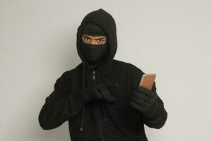 Portrait of mysterious man wearing black hoodie and mask doing hacking activity on mobile phone, hacker holding a smartphone. Isolated image on gray background photo