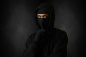 Mysterious man wearing black hoodie and mask standing against dark background, looking at camera. Dramatic low light portrait photo