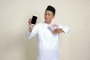 Portrait of attractive Asian muslim man in white shirt with skullcap showing one hundred thousand rupiah while showing blank screen mobile phone. Financial and shopping concept. Isolated image on gray photo