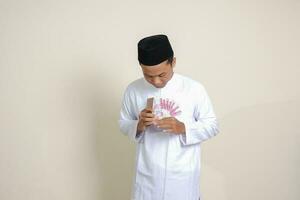 retrato de atractivo asiático musulmán hombre en blanco camisa demostración uno cien mil rupia mientras utilizando móvil teléfono. financiero y ahorros concepto. aislado imagen en gris antecedentes foto