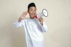 Portrait of attractive Asian muslim man in white shirt with skullcap speaking louder using megaphone, promoting product. Advertising concept. Isolated image on grey background photo