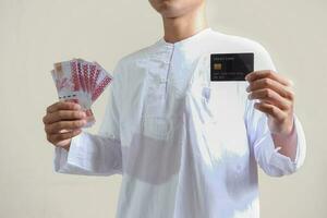 Portrait of attractive Asian muslim man in white shirt with skullcap holding one hundred thousand rupiah and presenting credit card. Isolated image on gray background photo