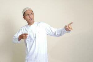 Portrait of attractive Asian muslim man in white shirt with skullcap showing product and pointing with his hand and finger to the side. Advertising concept. Isolated image on gray background photo
