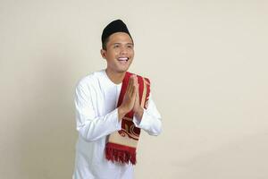 Portrait of attractive Asian muslim man in white shirt with skullcap showing apologize and welcome hand gesture. Isolated image on gray background photo