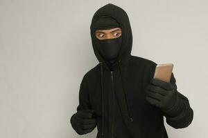 Portrait of mysterious man wearing black hoodie and mask doing hacking activity on mobile phone, hacker holding a smartphone. Isolated image on gray background photo