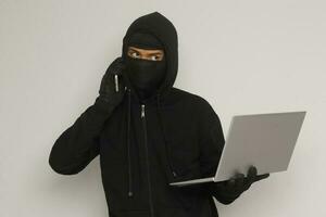 Portrait of mysterious man wearing black hoodie and mask doing hacking activity on laptop, hacker holding a mobile phone. Cyber security concept. Isolated image on gray background photo