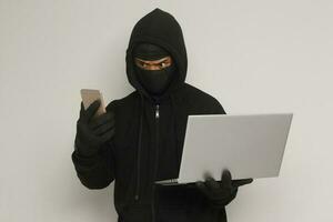 Portrait of mysterious man wearing black hoodie and mask doing hacking activity on laptop, hacker holding a mobile phone. Cyber security concept. Isolated image on gray background photo