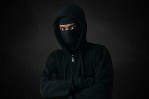 Mysterious man wearing black hoodie and mask standing against dark background, looking at camera. Dramatic low light portrait photo