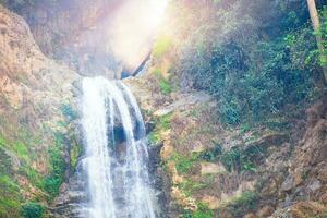 Beautiful waterfall in forestry with blue water with ripples on the surface. Blurred transparent blue colored clear calm water surface texture with splashes and bubbles. Water waves with shining. photo