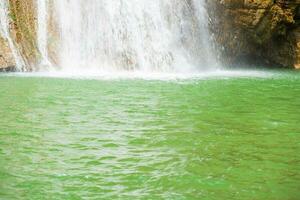 Beautiful waterfall in forestry with blue water with ripples on the surface. Blurred transparent blue colored clear calm water surface texture with splashes and bubbles. Water waves with shining. photo