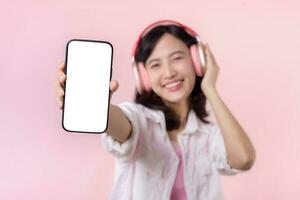 contento alegre sonriente asiático mujer con inalámbrico auriculares demostración blanco pantalla móvil teléfono o nuevo teléfono inteligente música solicitud anuncio Bosquejo aislado en rosado estudio antecedentes. foto