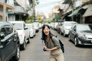 joven asiático mujer mochila viajero utilizando digital compacto cámara, disfrutando calle cultural local sitio y sonrisa. viajero comprobación fuera lado calles foto