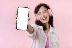 Happy cheerful smiling asian woman with wireless earphones showing blank screen mobile phone or new smartphone music application advertisement mockup isolated on pink studio background. photo