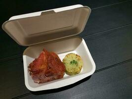 paper meal box filled with fried pork and cooked potato with spinach on black table with copy space photo