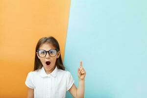 una niña de 7 años con gafas con cara de sorpresa apunta hacia la parte superior. educación infantil, concepto de aprendizaje. copie el espacio foto