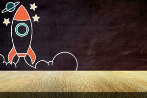 Wood Table with Retro Rocket in Chalk Drawing Style on Grunge Chalkboard Background, Suitable for Product Presentation Backdrop, Display, and Mock up in Back to School, Start up and Education Concept. photo