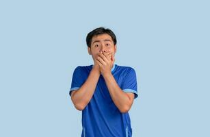 Portrait of excited young Asian man in blue shirt with hot news about sales discounts shocked covering mouth with hands isolated on blue background. People lifestyle concept photo