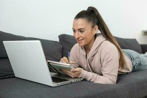 woman lying on the sofa at home taking notes in a notebook while looking at the laptop computer photo