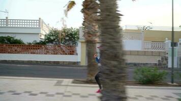 Woman with headphones runs down the street along the palm avenue at sunset. Healthy active lifestyle. Slow motion video