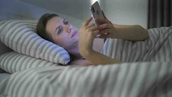 mujer usos un teléfono inteligente mientras acostado en cama. ella frota su ojos, porque ella es cansado y somnoliento. concepto de aumentado estrés y fatiga. video
