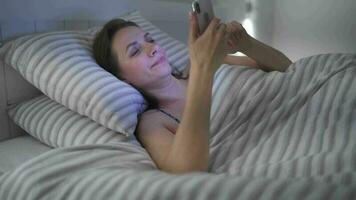 mujer usos un teléfono inteligente mientras acostado en cama. ella frota su ojos, porque ella es cansado y somnoliento. concepto de aumentado estrés y fatiga. video