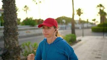 mujer con auriculares carreras abajo el calle a lo largo el palma avenida a puesta de sol. sano activo estilo de vida. lento movimiento video