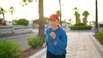 Woman with headphones runs down the street along the palm avenue at sunset. Healthy active lifestyle. Slow motion video