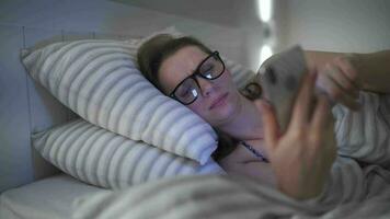 mulher usa uma Smartphone enquanto deitado dentro cama. ela esfrega dela olhos, Porque ela é cansado e com sono. conceito do aumentado estresse e fadiga. video