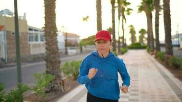 Woman with headphones runs down the street along the palm avenue at sunset. Healthy active lifestyle. Slow motion video