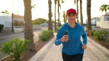 Woman with headphones runs down the street along the palm avenue at sunset. Healthy active lifestyle. Slow motion video